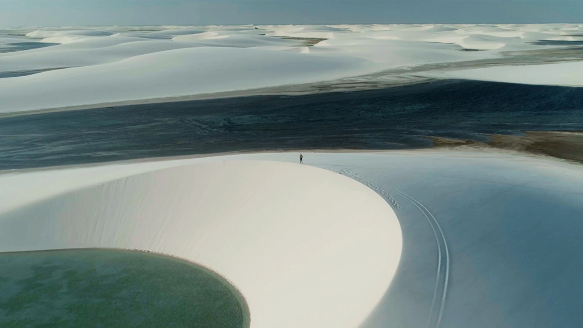 Aerial view of white dunes curving around a jade-green lagoon, one figure on a dune crest.